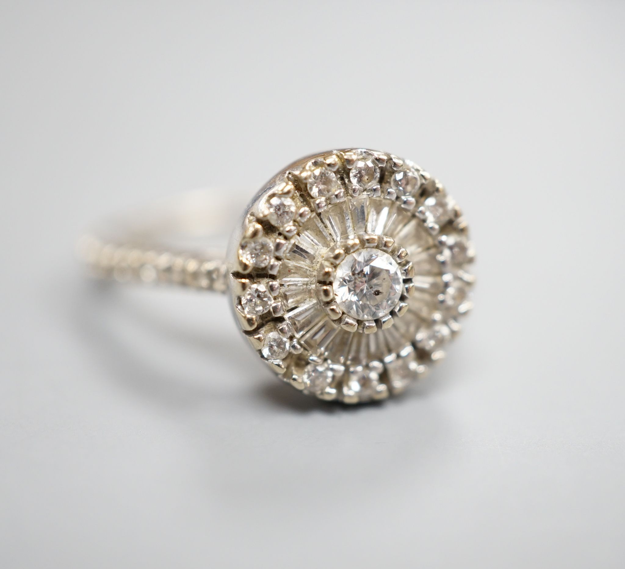 A modern white metal, round and trapeze cut diamond set target cluster ring, with diamond set shoulders, size N, gross weight 4.2 grams.